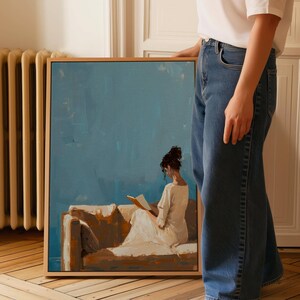 May include: A framed painting depicts a person reading on a sofa. The artwork features a woman in a white dress, seated on a tan sofa against a blue backdrop. The painting is in a wooden frame.