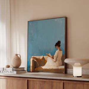 May include: A framed painting depicts a person reading on a sofa, set against a blue backdrop. The artwork is in a gold frame and is displayed on a wooden cabinet. A beige vase, books, and a lamp are also on the cabinet.
