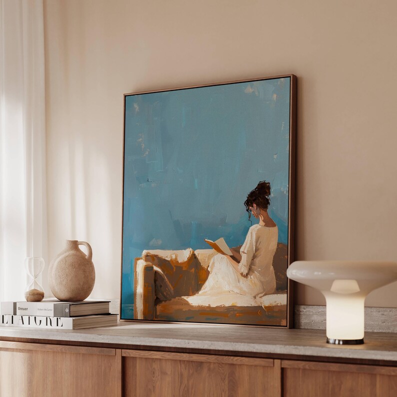 May include: A framed painting depicts a person reading on a sofa, set against a blue backdrop. The artwork is in a gold frame and is displayed on a wooden cabinet. A beige vase, books, and a lamp are also on the cabinet.