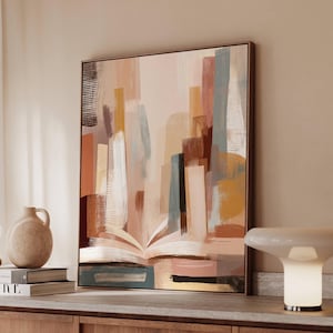 May include: Abstract art print featuring a cityscape design in muted tones of beige, brown, and blue. The artwork depicts an open book with buildings rising from its pages. The framed canvas is displayed on a wooden cabinet, with a lamp and vase nearby.