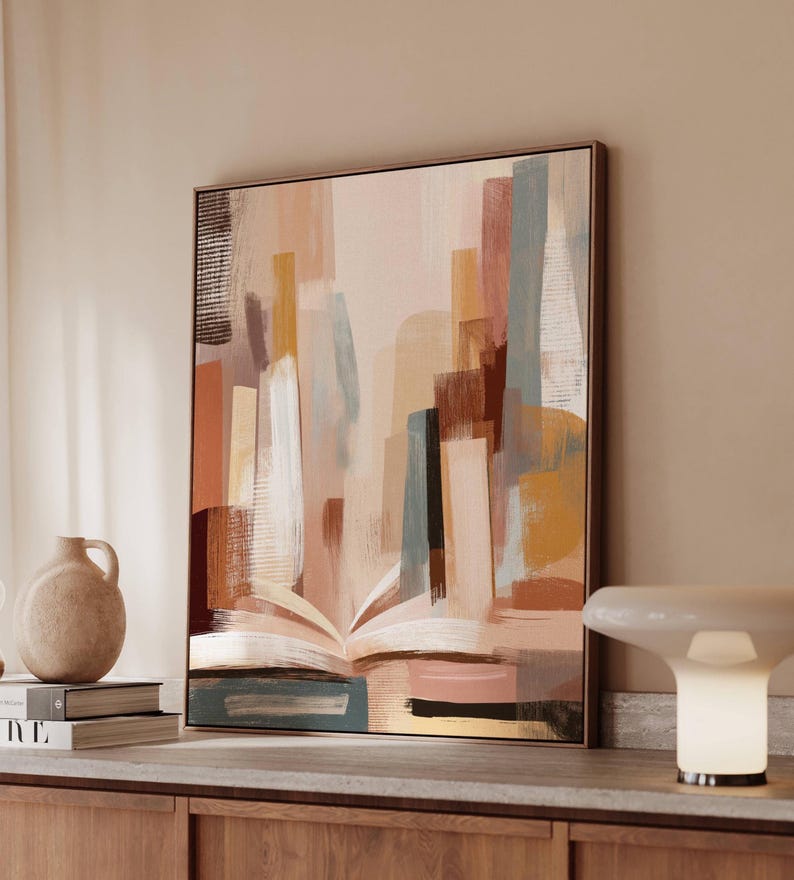 May include: Abstract art print featuring a cityscape design in muted tones of beige, brown, and blue. The artwork depicts an open book with buildings rising from its pages. The framed canvas is displayed on a wooden cabinet, with a lamp and vase nearby.