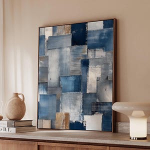 May include: Abstract art piece featuring a patchwork design in shades of blue, white, and beige. The artwork is framed in a brown wooden frame and hangs on a beige wall. The painting has a textured appearance.
