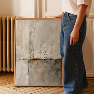 May include: Abstract art piece in a wooden frame. The artwork features a textured surface with a palette of gray, white, and brown tones. The composition includes vertical and horizontal elements, creating a layered effect. The artwork is displayed indoors.