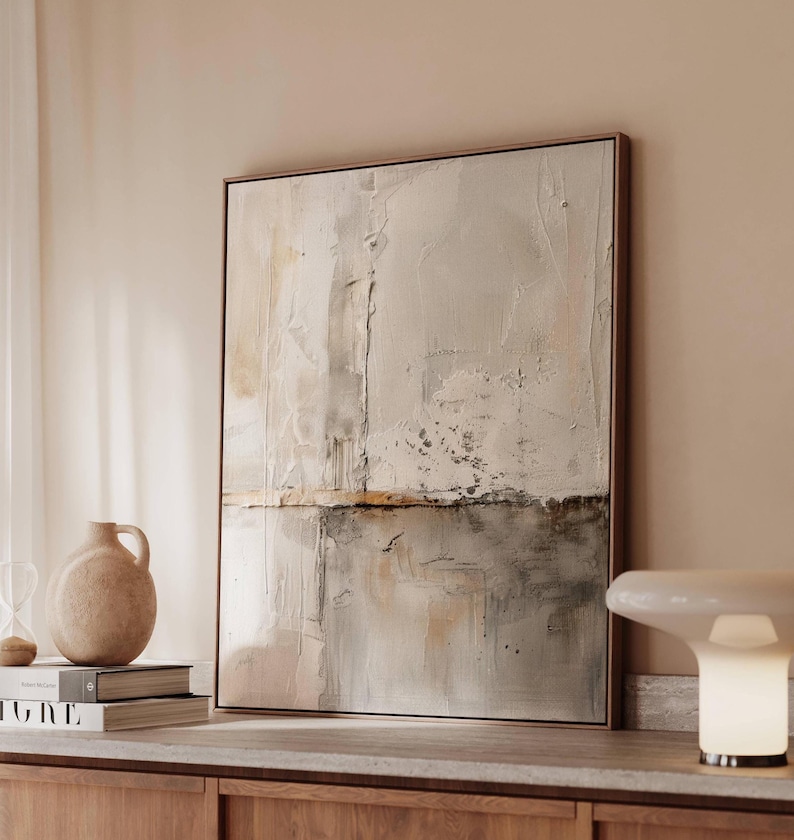 May include: Abstract art piece in a wooden frame. The artwork features a textured, neutral color palette with shades of beige, gray, and brown. The composition is divided horizontally, with a lighter upper section and a darker lower section.
