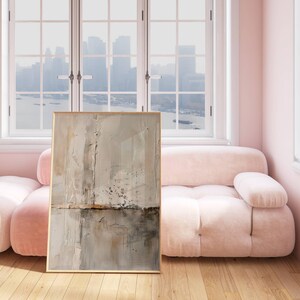 May include: Abstract art print in a gold frame. The artwork features a textured, neutral color palette with shades of beige, gray, and brown. The painting is displayed in a room with a pink sofa and a large window overlooking a cityscape.