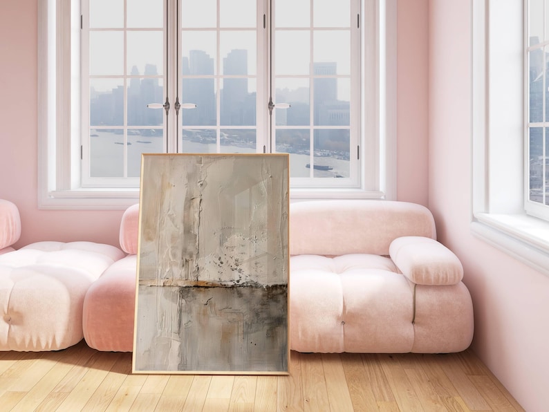May include: Abstract art print in a gold frame. The artwork features a textured, neutral color palette with shades of beige, gray, and brown. The painting is displayed in a room with a pink sofa and a large window overlooking a cityscape.