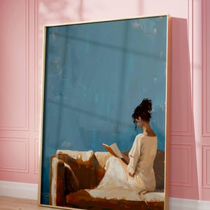 May include: An oil painting depicts a person reading on a brown sofa against a blue backdrop. The person is wearing a white dress. The artwork is framed in a gold frame and leans against a pink wall.