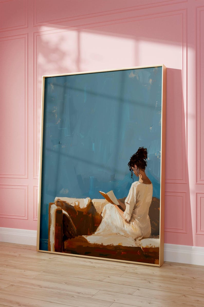 May include: An oil painting depicts a person reading on a brown sofa against a blue backdrop. The person is wearing a white dress. The artwork is framed in a gold frame and leans against a pink wall.