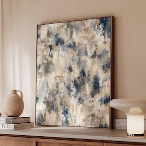 May include: Abstract art print in a wooden frame. The artwork features a textured design with brushstrokes in shades of beige, blue, and gray. The framed art is displayed on a wooden cabinet, alongside a ceramic vase and books.