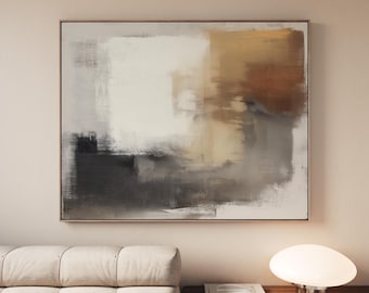 May include: Abstract art painting with a gold frame. The artwork features a blend of neutral colors, including white, gray, and brown, creating a textured, layered effect. The painting is displayed above a beige sofa and a wooden nightstand with a lamp.
