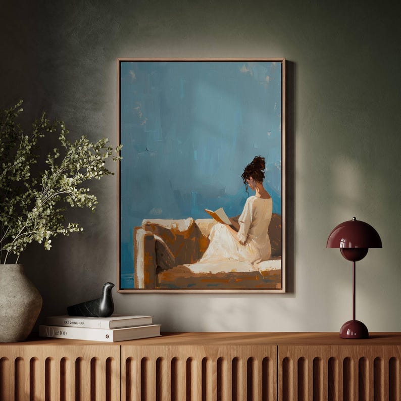 May include: A framed painting depicts a person reading on a sofa against a blue backdrop. The artwork is in a wooden frame and hangs above a wooden cabinet. A burgundy lamp and decorative items are on the cabinet.
