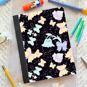 May include: A black notebook with a colorful butterfly pattern. The butterflies are in shades of yellow, purple, and blue. The notebook is surrounded by art supplies, including pencils, a paintbrush, and scissors.