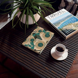 May include: A decorative notebook with a teal and beige pattern featuring keys, books, and circles, with the word "Love" on the cover. A "Travel Magazine" and a cup of coffee sit nearby on a dark wooden table.