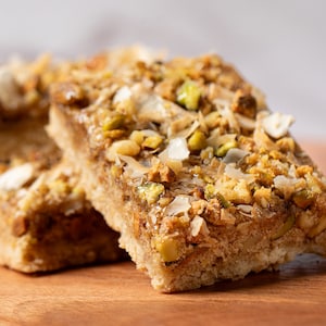 May include: Close-up of a stack of three square-shaped bars topped with a mixture of chopped pistachios, shredded coconut, and a light brown glaze. The bars are sitting on a wooden surface.