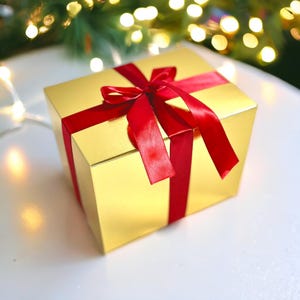 May include: A gold gift box with a red satin ribbon and bow. The box is sitting on a white surface.