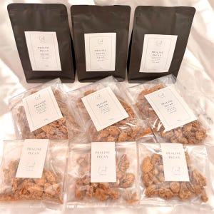 May include: An assortment of pecan pralines in clear plastic bags and dark brown paper bags. Each bag has a white label with the text "PRALINE PECAN" and the brand's logo. The pralines appear to be coated in a sugary glaze.