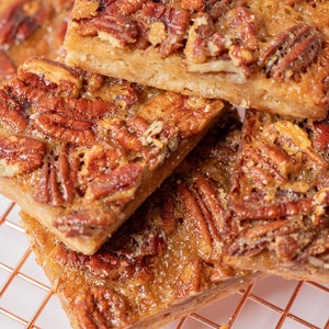 May include: A close-up of pecan pie bars stacked on a copper wire rack. The bars are golden brown and have a glossy finish. The pecans are evenly distributed throughout the bars.