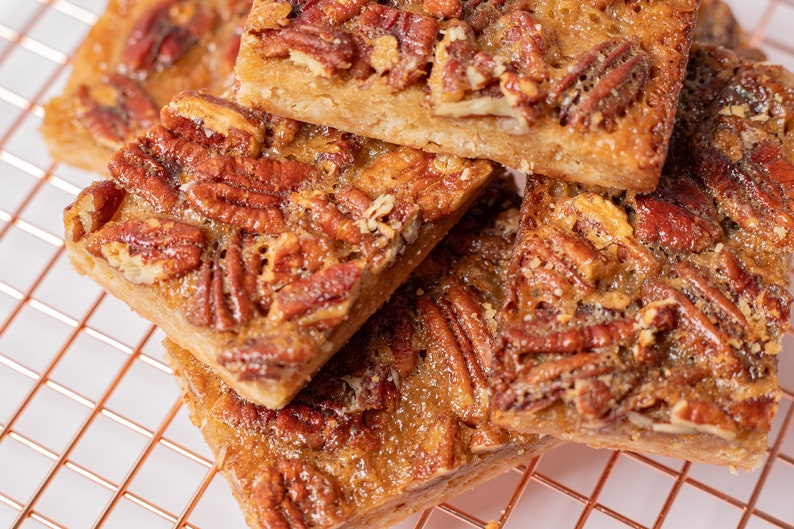 May include: A close-up of pecan pie bars stacked on a copper wire rack. The bars are golden brown and have a glossy finish. The pecans are evenly distributed throughout the bars.