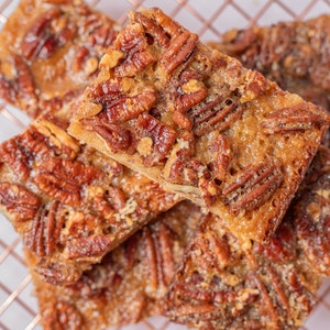 May include: A stack of four pecan bars, each with a golden brown caramel topping and pecans, on a copper wire rack.