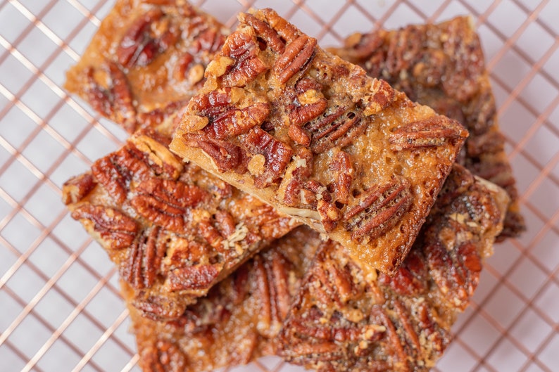 May include: A stack of four pecan bars, each with a golden brown caramel topping and pecans, on a copper wire rack.