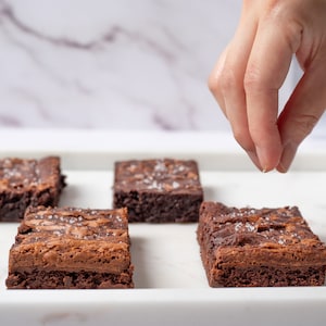 May include: A white rectangular plate with six chocolate brownies topped with sea salt. A hand is sprinkling more salt on one of the brownies.
