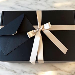 May include: A black gift box with a white satin ribbon tied in a bow. A black envelope is placed on top of the box.