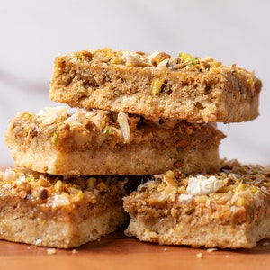 May include: A stack of four square-shaped shortbread cookies topped with chopped pistachios and white coconut flakes. The cookies are arranged on a wooden surface.