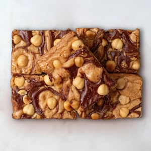 May include: A close-up of a stack of six chocolate and hazelnut swirl brownies with whole hazelnuts on top. The brownies are on a white marble surface.