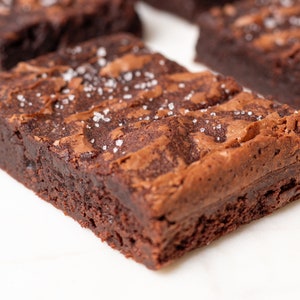 May include: A close-up of a chocolate brownie with a flaky, fudgy texture. The brownie is sprinkled with coarse salt crystals.