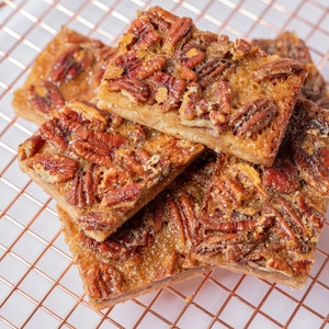 A delicious pecan bar featuring a buttery shortbread cookie crust, topped with a gooey, crunchy layer of caramelized pecans for the perfect balance of sweet and rich flavors.