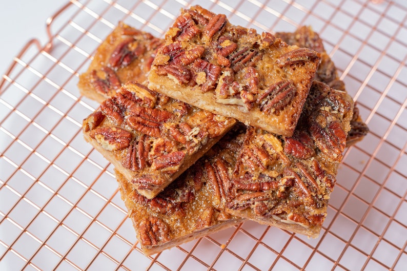 A delicious pecan bar featuring a buttery shortbread cookie crust, topped with a gooey, crunchy layer of caramelized pecans for the perfect balance of sweet and rich flavors.