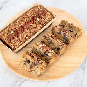 May include: A loaf of pecan bread in a cardboard container and several slices of bread on a wooden plate. The bread is topped with pecans and a crumbly topping. The bread slices are approximately 4 inches long.