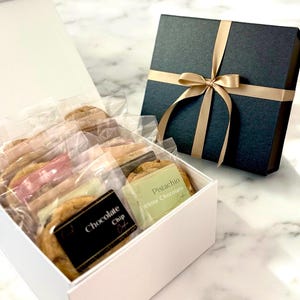 May include: A white gift box filled with individually wrapped cookies, including chocolate chip and pistachio white chocolate varieties. A separate navy blue gift box with a gold ribbon is also present.