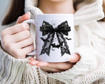 May include: White ceramic mug with a black lace bow and bats design. The bow is tied with a black ribbon and has a spiderweb pattern.