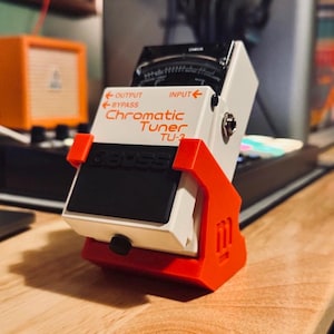 May include: A white and black BOSS Chromatic Tuner TU-2, with red accents, is mounted in a red holder. The tuner has a display and input/output jacks. An orange amplifier is visible in the background.