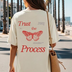 Op de afbeelding: Crèmekleurig T-shirt met de rode tekst "Trust the Process" en een rode vlinder. Het shirt heeft een losse pasvorm en wordt op een zonnige dag buiten gedragen.