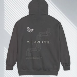 Puede incluir: Una sudadera con capucha gris oscuro con un gráfico de mariposa blanca y el texto "WE ARE ONE" impreso en la espalda. La sudadera también presenta un diseño abstracto blanco y una pequeña cita.