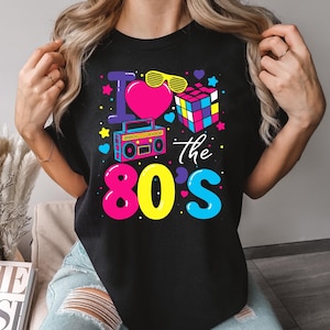 K&ouml;nnte beinhalten: Schwarzes T-Shirt mit einem farbenfrohen Aufdruck "I love the 80s". Das Design umfasst ein pinkes Herz, gelbe Sonnenbrillen, einen Ghettoblaster, einen Rubik's Cube und Sterne. Das Wort "BLACK" ist unten rechts aufgedruckt.