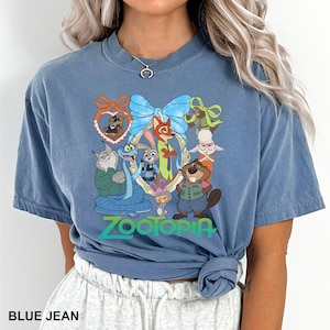 K&ouml;nnte beinhalten: Blaues Jeans-T-Shirt mit einer farbenfrohen Grafik von Charakteren aus dem Animationsfilm Zootopia. Die Grafik zeigt verschiedene Tierfiguren in einem dekorativen Rahmen, mit dem Wort "Zootopia" in einem Farbverlauf.