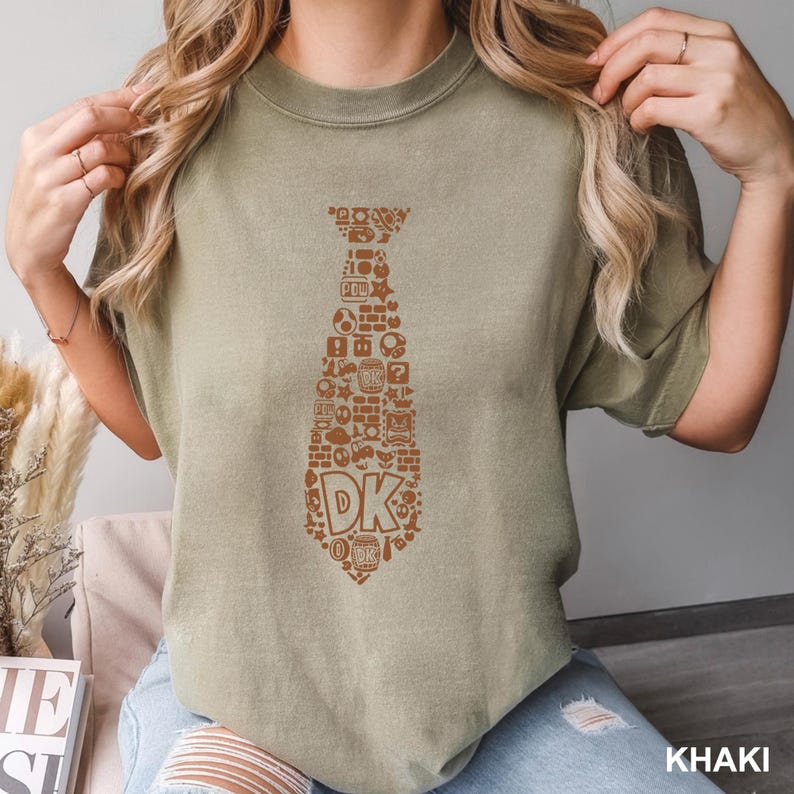 May include: Khaki-colored t-shirt featuring a brown tie design filled with video game-themed icons and the letters "DK". The shirt has short sleeves and a relaxed fit. The word "KHAKI" is printed in the bottom right corner.