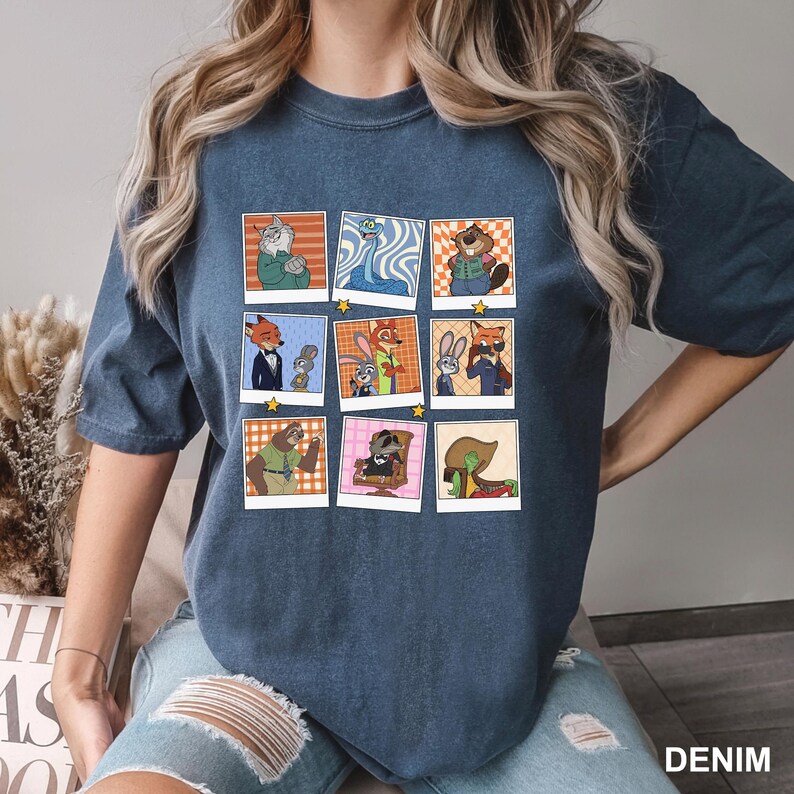 May include: Denim blue t-shirt featuring a grid of cartoon character portraits. Each portrait is framed in a square with a unique background and star accents. The shirt's fabric has a slightly faded appearance.