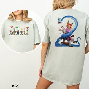 Op de afbeelding: Lichtgroen T-shirt met cartoon illustraties op de voor- en achterkant. De voorkant toont cartoonfiguren met hartvormige ballonnen. De achterkant heeft een blauwe slang met cartoonfiguren.