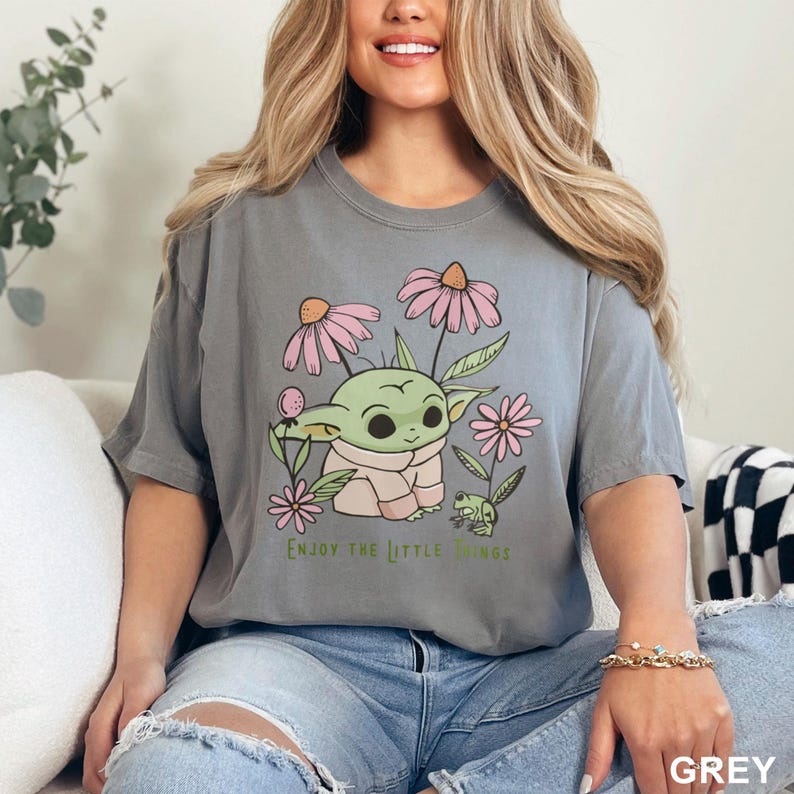 Puede incluir: Camiseta gris con un personaje de dibujos animados verde rodeado de flores rosas y una peque&ntilde;a rana verde. El texto "ENJOY THE LITTLE THINGS" est&aacute; impreso debajo. La camiseta es de material suave.