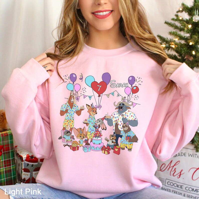 May include: Light pink sweatshirt featuring a colorful graphic of Zootopia characters celebrating a birthday. The design includes balloons, a cake, and the name "Sarah" with the number "7". The sweatshirt is a crew neck style.