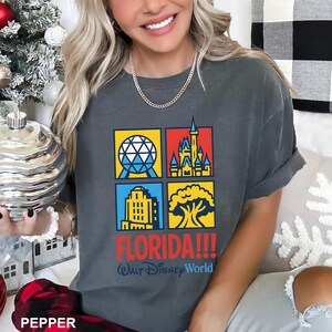 Może przedstawiać: Ciemnoszara koszulka z czterema kolorowymi kwadratami przedstawiającymi kultowe obrazy Florydy. Koszulka ma napis "FLORIDA!!!" w kolorze czerwonym i "Walt Disney World" w kolorze niebieskim. Widoczny srebrny łańcuszek.