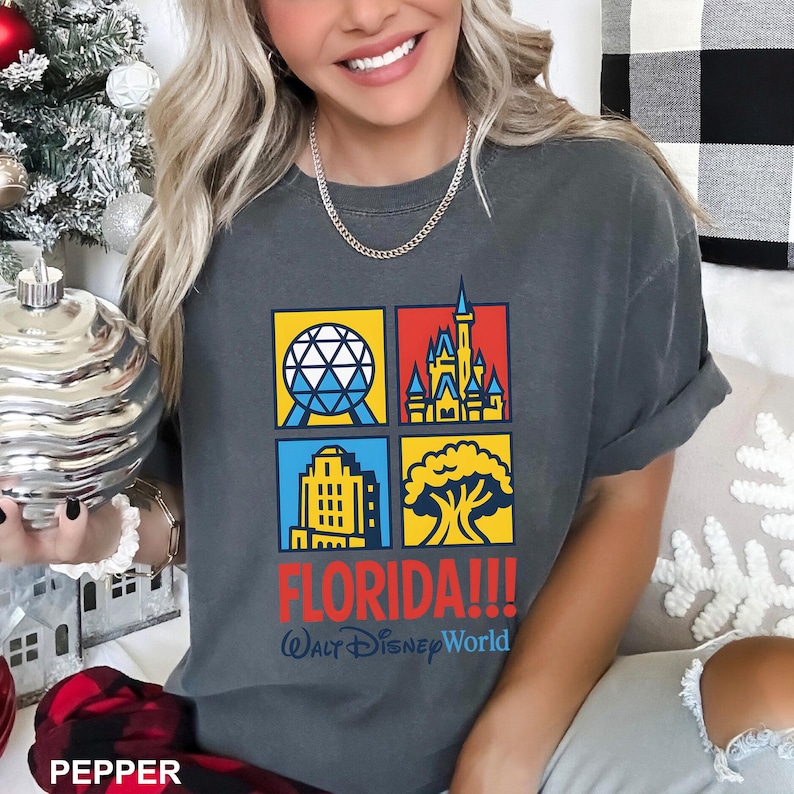 Może przedstawiać: Ciemnoszara koszulka z czterema kolorowymi kwadratami przedstawiającymi kultowe obrazy Florydy. Koszulka ma napis "FLORIDA!!!" w kolorze czerwonym i "Walt Disney World" w kolorze niebieskim. Widoczny srebrny łańcuszek.