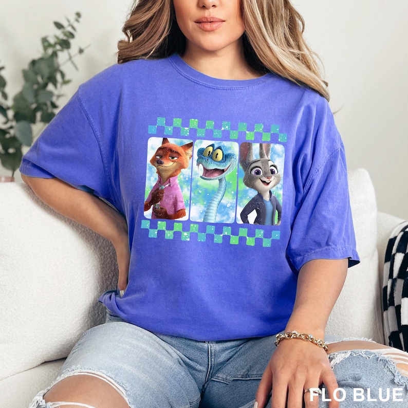 May include: A blue t-shirt featuring a graphic of three cartoon characters: a fox, a snake, and a rabbit. The graphic is framed with a green and blue checkered pattern. The text "FLO BLUE" is visible at the bottom right.