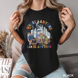 May include: Black t-shirt with a colorful graphic featuring cartoon animals and the text "WHERE ANYONE CAN BE ANYTHING". The design includes a cityscape and various animal characters.