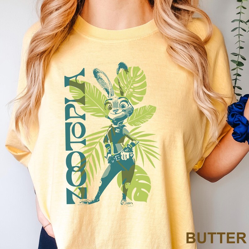 May include: Yellow t-shirt featuring the word "Zootopia" in green vertical text. The shirt has a graphic of Judy Hopps, a rabbit character, with green leaves in the background. The word "BUTTER" is printed at the bottom right.