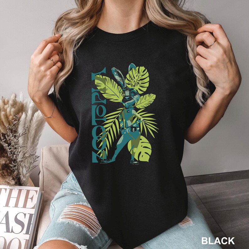 May include: Black t-shirt featuring a graphic of a rabbit-like figure in a futuristic outfit, surrounded by green tropical leaves. The word "TOPPY" is printed vertically in teal. The word "BLACK" is printed in white at the bottom.